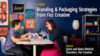 Fizz Creative founders, Jason and Katie Melnick are in a room seated opposite each other on a white table looking at designs, a white lamp and shelf to the right. The purple wall is decorated with varying sizes of typography and design.